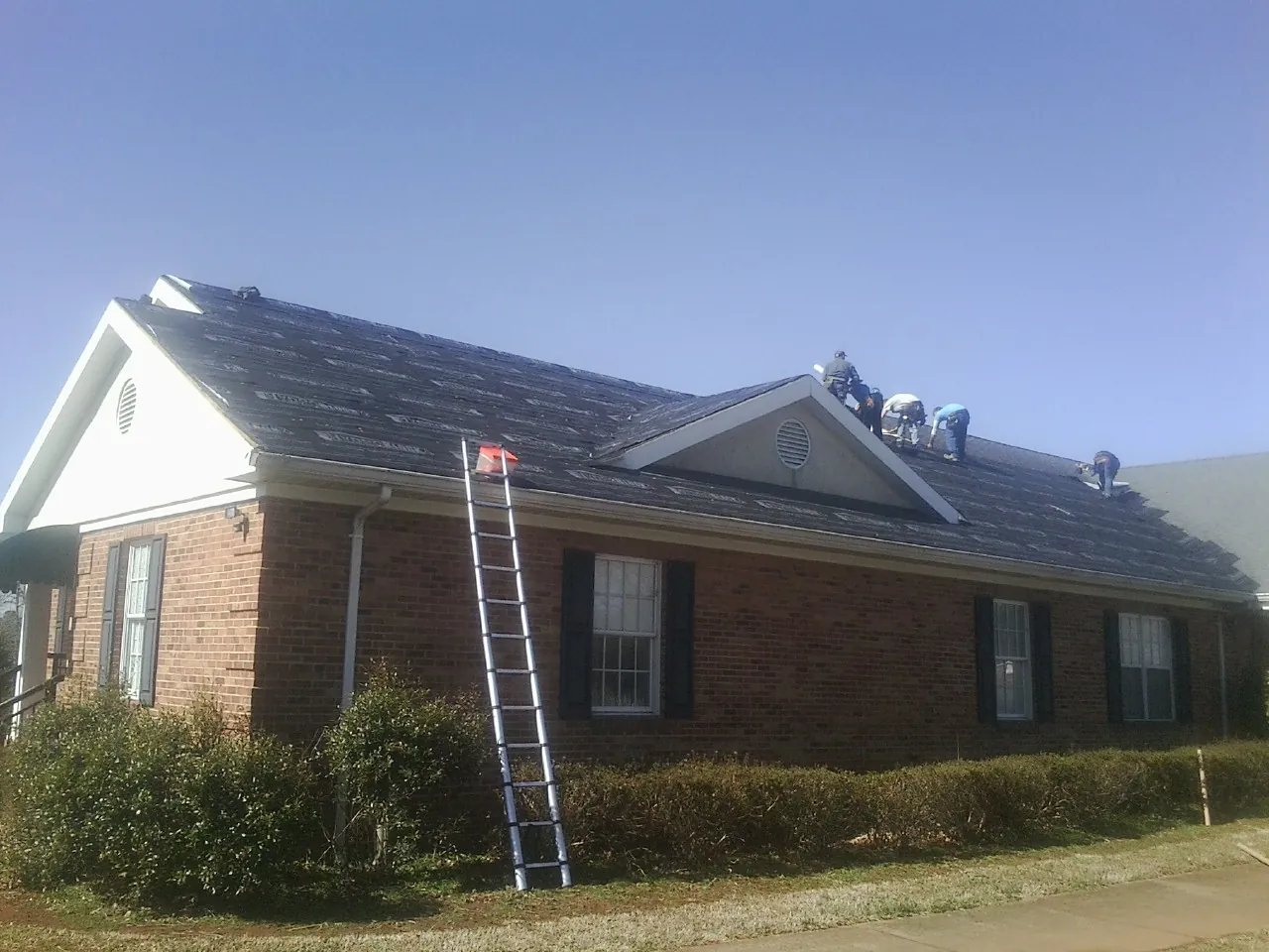 Crew tearing shingles off a brick ranch roof with a ladder set — Metal Roof Repair work by our crew in Phelps
