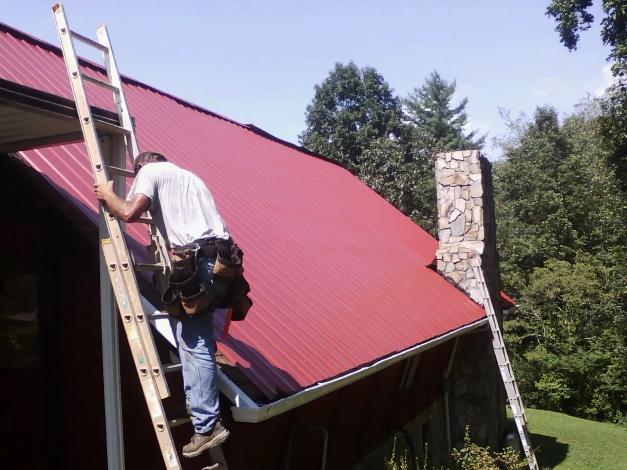 Roofer on a ladder beside a red metal roof and stone chimney — Soffit & Fascia Repair by Roofing Brothers in Phelps